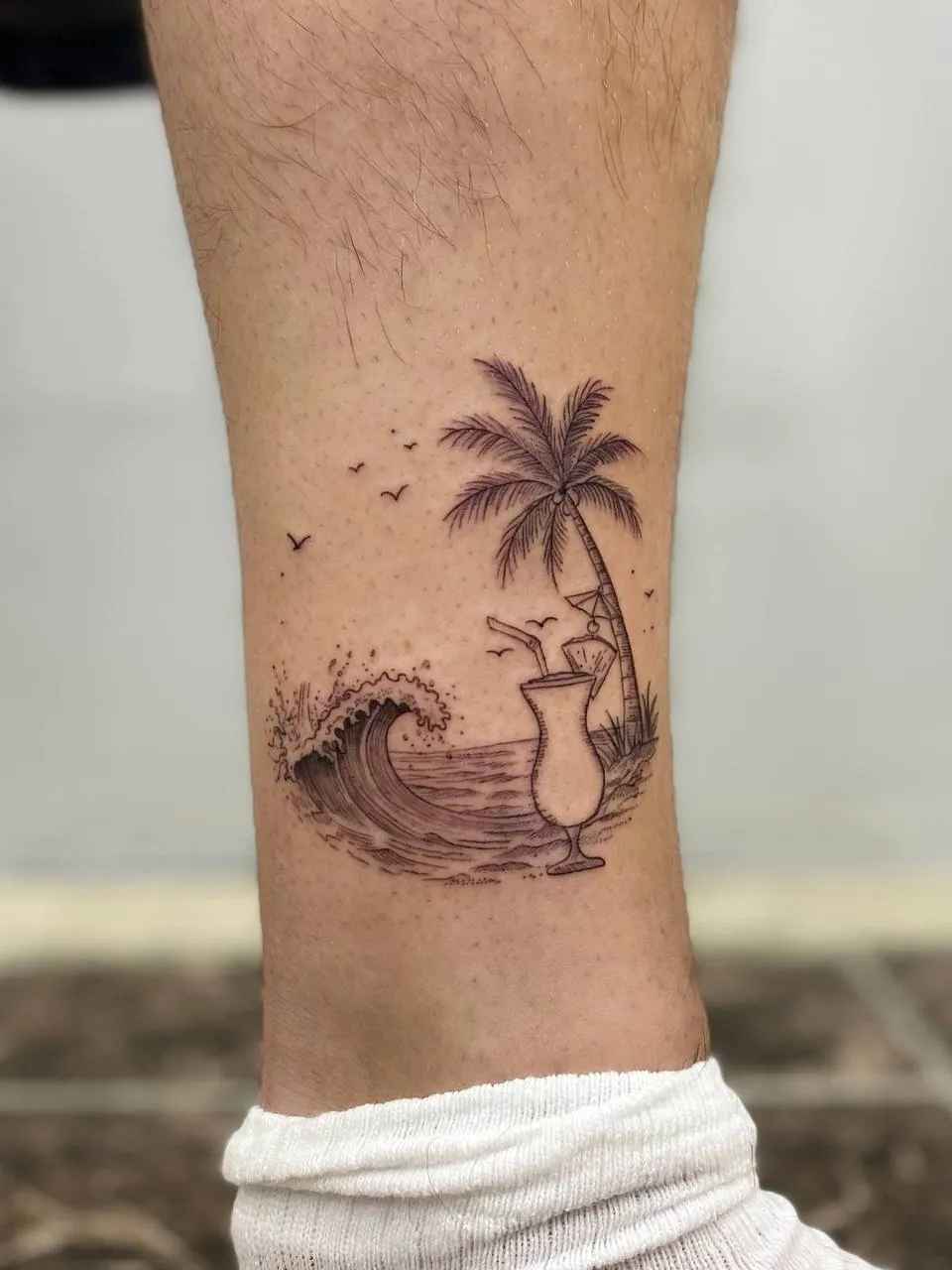 Fine-line tattoo of a palm tree, a breaking wave and a cocktail glass on the lower leg