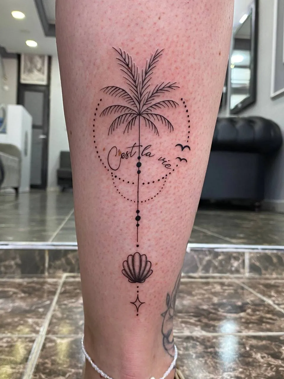 Fine-line palm tree in a dotted circle with the words C’est la vie and a seashell on the calf